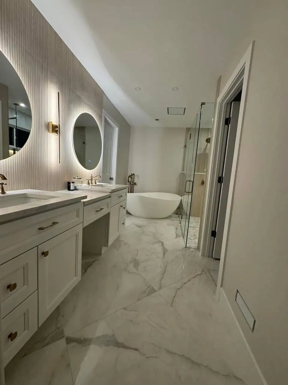 Master bathroom remodel by Devlin Remodeling, home remodeler in Independence, KY: freestanding tub, backlit round mirrors, white shaker vanity and marble-look floor